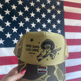 Load image into Gallery viewer, Olive Green and Green Camo Trucker Hat with more guns less crime Trump 2024. Graphic of A Large Hat on Donald Trump holding a shotgun.