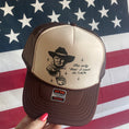 Load image into Gallery viewer, A brown trucker hat with a graphic print of a character, text, and a sparkling water bottle, set against a red and white fabric background.Hat says "The Only Joe I need at 7am."