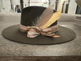 Load image into Gallery viewer, A black Flatbrim hat with 2 large feathers one pink and one gold, then tied in perfect rows are blush pink ribbons and silks and a Carmel studded gold cord.