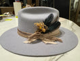 Load image into Gallery viewer, Silver flat brim hat with gold hues of silk and cord and dried florals and deep blue feathers to complete this up close styled hat at a private event.