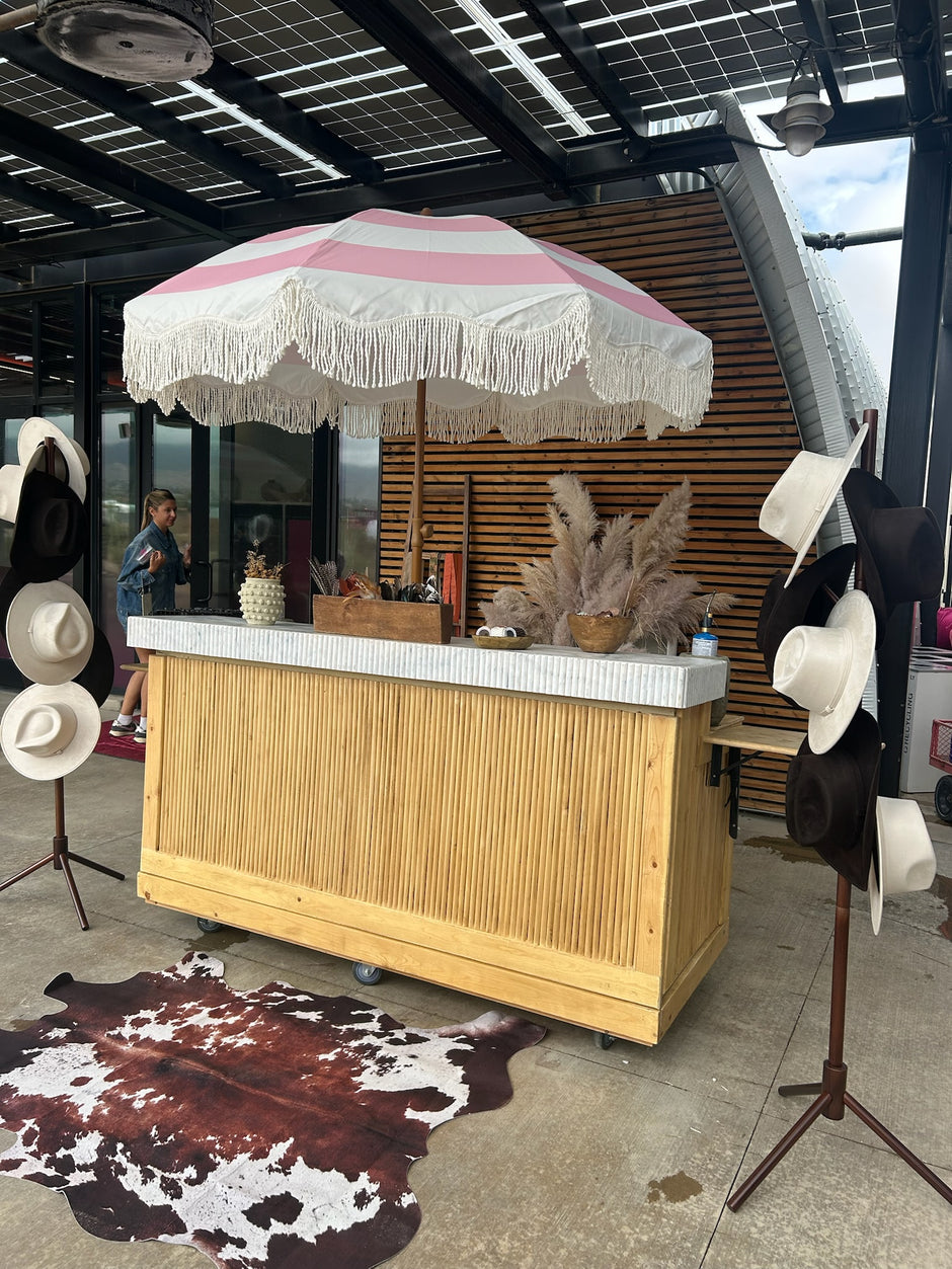 Wooden bar with pink and white striped umbrella and cowboy hats on stands in an outdoor setting.