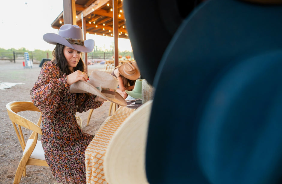 Woman in a patterned dress and cowboy hat sitting at a table outdoors with a blurred foreground. She has a Beautiful Signature Hat They are styling together.