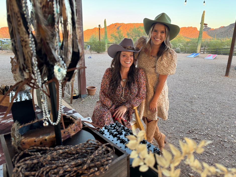Two women standing outdoors with a scenic desert background. Infront of the two women is their branding and Bolo Tie Bar Set Up to have a Cooperate event and Fun Night Out Making Bolo ties and Branding Hats.